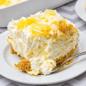 May include: A square slice of pineapple coconut dessert on a white plate with a silver fork. The dessert features a golden brown crumb base, a thick layer of white cream, topped with bright yellow pineapple chunks and shredded coconut flakes.