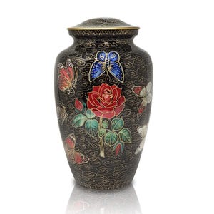Puede incluir: Urna negra con tapa, adornada con una rosa roja, mariposas y detalles dorados. La urna decorativa presenta un diseño floral y de mariposas, adecuada para fines conmemorativos.
