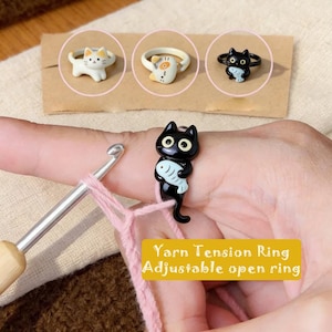 Peut inclure: Une bague ouverte réglable représentant un chat noir tenant un petit poisson, utilisée comme guide de tension de fil pour le crochet. La bague est portée sur un doigt avec un crochet en métal et du fil rose. Texte : Yarn Tension Ring Adjustable open ring.