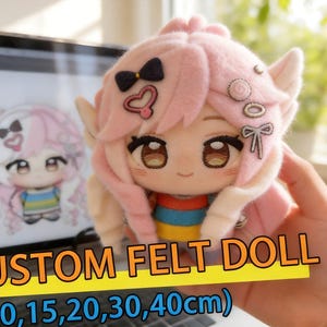 Puede incluir: Una muñeca de peluche con cabello rosa claro, ojos bordados marrones y una pequeña sonrisa. La muñeca presenta accesorios de fieltro para el cabello, incluyendo un lazo negro, un corazón rosa y un lazo blanco. Viste una camisa con rayas horizontales rojas, azules y amarillas.
