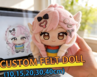 Puede incluir: Una muñeca de peluche con cabello rosa claro, ojos bordados marrones y una pequeña sonrisa. La muñeca presenta accesorios de fieltro para el cabello, incluyendo un lazo negro, un corazón rosa y un lazo blanco. Viste una camisa con rayas horizontales rojas, azules y amarillas.