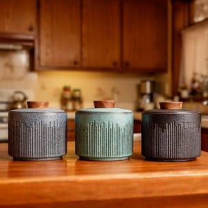 Peut inclure: Trois bocaux en céramique avec couvercles en bois. Les bocaux sont dans des tons de bleu, de vert d'eau et de noir. Chaque bocal a un motif texturé à lignes verticales. Les couvercles sont en bois clair. Les bocaux sont sur une surface en bois.