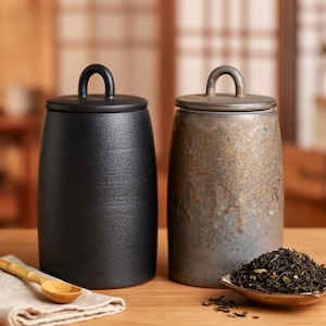 Può includere: Due barattoli in ceramica con coperchi. Uno è nero opaco, l'altro è grigio e oro screziato. Un piccolo cucchiaio di legno poggia su un tovagliolo di lino davanti al barattolo nero. Un piattino di foglie di tè è davanti al barattolo grigio.