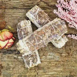 Könnte beinhalten: Drei Bündel getrockneten Salbeis, mit weißem Faden gebunden, auf einer verwitterten Holzoberfläche. Eine getrocknete Rose mit roten und gelben Blütenblättern und ein Zweig rosa Blüten sind ebenfalls vorhanden.