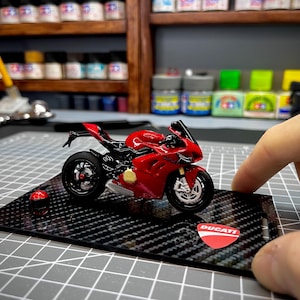 Könnte beinhalten: Rotes Ducati Motorradmodell mit schwarzen Akzenten, präsentiert auf einer Basis im Carbonfaser-Stil. Das Modell verfügt über detaillierte Designelemente und das Ducati-Logo. Ein kleiner roter Helm ist ebenfalls vorhanden.