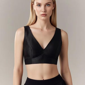 May include: A black sleeveless crop top with a deep V-neck. The top has a textured appearance and is paired with a black skirt. The model has blonde hair and is looking directly at the camera.