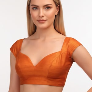 Puede incluir: Un top corto naranja quemado con escote cuadrado y mangas cortas. El top tiene un escote en V en el centro y está hecho de una tela texturizada. La prenda es de color liso y parece ser una blusa.