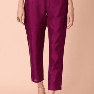 May include: A pair of plum-colored, tailored pants. The pants have a high waist and a straight leg, ending just above the ankles. The fabric has a subtle sheen. The model is wearing gold-colored heels.