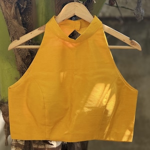 May include: Mustard yellow halter top with a high neckline and a keyhole back detail. The sleeveless blouse is hanging on a wooden hanger. The fabric has a subtle sheen.