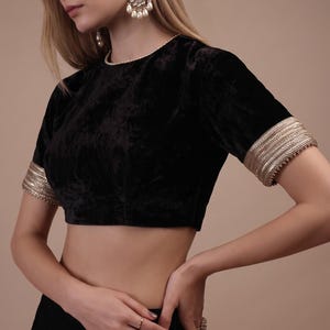May include: A black velvet blouse featuring a round neckline and short sleeves with wide, metallic gold embroidered cuffs. The garment is paired with a matching black skirt adorned with small, repeating floral embroidery patterns in silver and pink.