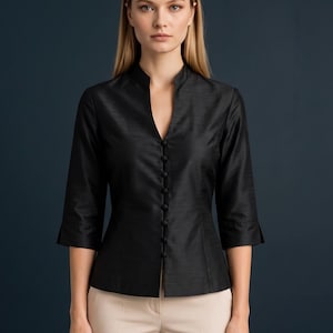May include: A black blouse with a mandarin collar, a deep V-neckline, and a front button placket featuring small fabric-covered buttons. The garment has three-quarter length sleeves with subtle flared cuffs and a textured, slightly lustrous finish.