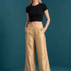 Könnte beinhalten: Ein schwarzes kurzärmeliges bauchfreies Oberteil kombiniert mit einer goldfarbenen Hose mit weitem Bein. Die Hose hat eine hohe Taille mit Seitentaschen und eine leicht strukturierte Oberfläche. Das Outfit wird durch goldene Sandalen mit Blockabsatz ergänzt.