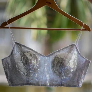 Puede incluir: Un top plateado con lentejuelas y tirantes finos cuelga de una percha de madera. El top tiene un escote en V y está cubierto de lentejuelas brillantes. El diseño presenta un área de busto estructurada.