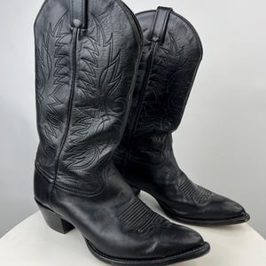 May include: A pair of black leather cowboy boots featuring a pointed toe, a stacked heel, and decorative tonal stitching patterns on the shaft and vamp. The boots have pull tabs at the top of the shaft and a smooth finish throughout the exterior.