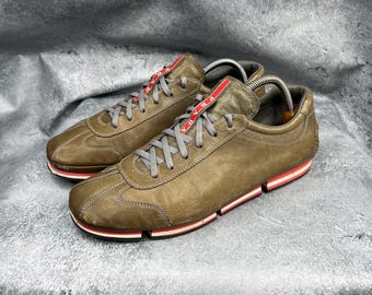 May include: Olive green sneakers with grey laces and red detailing. The low-profile shoes have a stitched upper and a red and white striped sole. The tongue features a red tag.