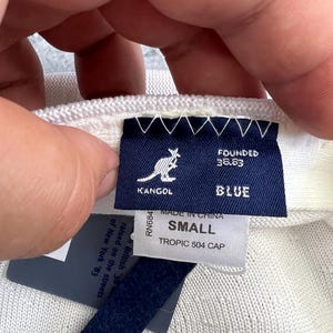 Puede incluir: Primer plano de una gorra Kangol Tropic 504 blanca. La gorra tiene una etiqueta azul con el logotipo de Kangol, la palabra "BLUE" y el texto "SMALL". La etiqueta tambi&eacute;n dice "MADE IN CHINA" y "TROPIC 504 CAP".