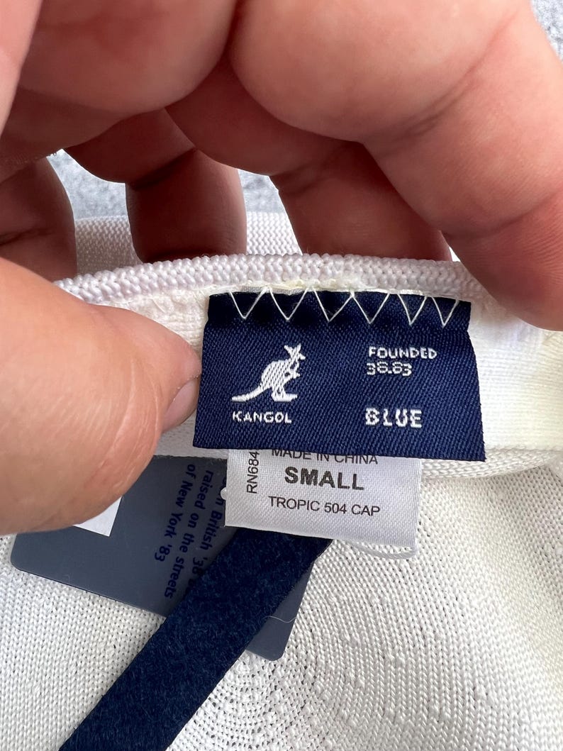 Puede incluir: Primer plano de una gorra Kangol Tropic 504 blanca. La gorra tiene una etiqueta azul con el logotipo de Kangol, la palabra "BLUE" y el texto "SMALL". La etiqueta tambi&eacute;n dice "MADE IN CHINA" y "TROPIC 504 CAP".