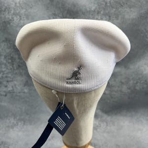 Puede incluir: Un gorro Kangol blanco con un logotipo plateado. El sombrero se exhibe en una cabeza de maniqu&iacute; con una etiqueta adjunta. El sombrero est&aacute; hecho de una tela texturizada.