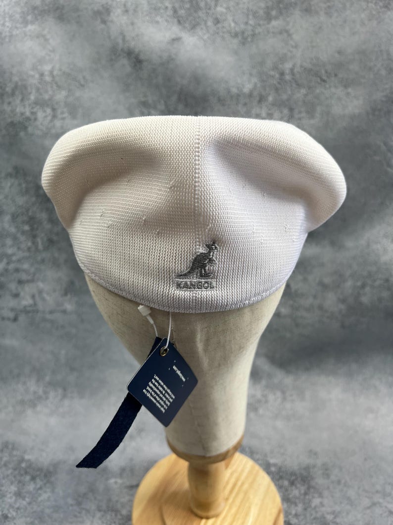 Puede incluir: Un gorro Kangol blanco con un logotipo plateado. El sombrero se exhibe en una cabeza de maniqu&iacute; con una etiqueta adjunta. El sombrero est&aacute; hecho de una tela texturizada.