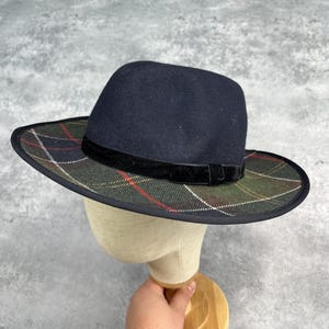 May include: A navy blue fedora hat with a plaid brim in green, red, and white. The hat has a black velvet band and is displayed on a mannequin head. The hat's brim measures approximately 7.6 cm.