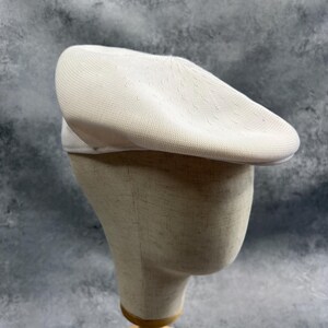 Puede incluir: Un gorro de boina beige claro hecho de tejido de punto. El sombrero se exhibe en una cabeza de maniqu&iacute; con una base de madera. La boina tiene una corona redondeada y un ala peque&ntilde;a. El fondo es gris moteado.