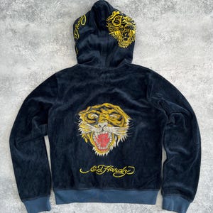May include: Navy blue hooded sweatshirt with a tiger design on the back and hood. The tiger is yellow and gold with red accents. The sweatshirt has the text "Ed Hardy" in yellow script.