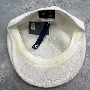Puede incluir: Gorra Kangol Tropic 504 blanca con un dise&ntilde;o de punto texturizado. La gorra tiene una peque&ntilde;a visera y una etiqueta que dice "BLUE" y "TROPIC 504 CAP". La gorra es de talla peque&ntilde;a.