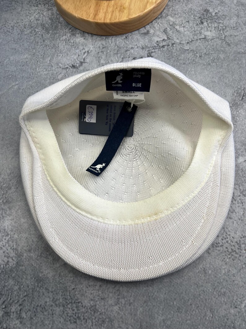 Puede incluir: Gorra Kangol Tropic 504 blanca con un dise&ntilde;o de punto texturizado. La gorra tiene una peque&ntilde;a visera y una etiqueta que dice "BLUE" y "TROPIC 504 CAP". La gorra es de talla peque&ntilde;a.