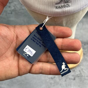 Puede incluir: Gorra Kangol gris claro con el logotipo de la marca bordado en el lateral. La etiqueta adjunta muestra el logotipo de Kangol y el texto "Born British, raised on the streets of New York '83."