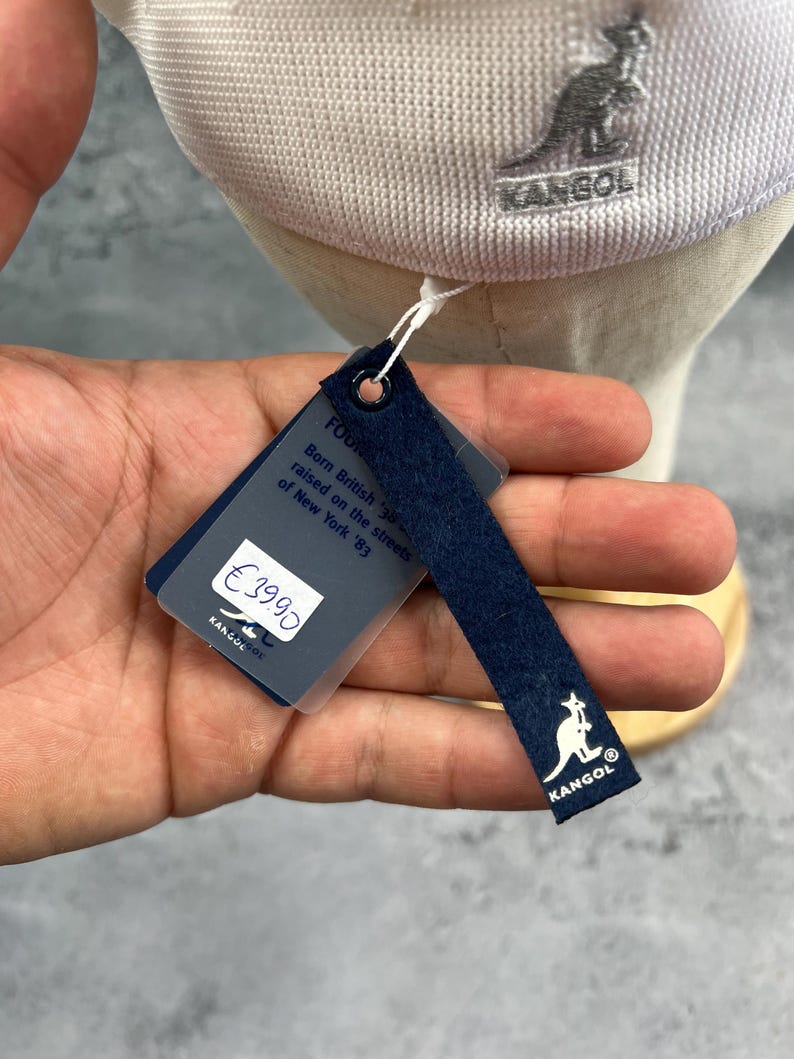 Puede incluir: Gorra Kangol gris claro con el logotipo de la marca bordado en el lateral. La etiqueta adjunta muestra el logotipo de Kangol y el texto "Born British, raised on the streets of New York '83."