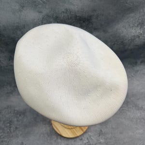 Puede incluir: Un gorro de boina blanco tejido, exhibido sobre un soporte de madera. El sombrero tiene un dise&ntilde;o circular texturizado y una forma suave y redondeada. La boina es de color neutro, adecuada para varios atuendos.