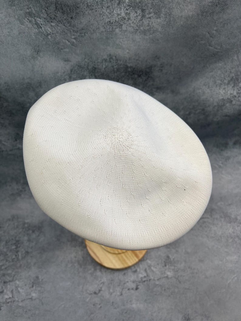 Puede incluir: Un gorro de boina blanco tejido, exhibido sobre un soporte de madera. El sombrero tiene un dise&ntilde;o circular texturizado y una forma suave y redondeada. La boina es de color neutro, adecuada para varios atuendos.