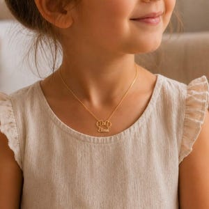 Op de afbeelding: Een goudkleurige ketting met een hanger in de vorm van een strik en de naam "Elena" in een sierlijk schrift. De ketting wordt gedragen door een persoon met een lichtgekleurde top.