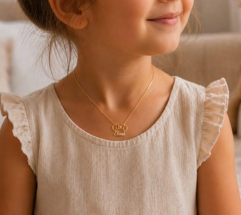 Op de afbeelding: Een goudkleurige ketting met een hanger in de vorm van een strik en de naam "Elena" in een sierlijk schrift. De ketting wordt gedragen door een persoon met een lichtgekleurde top.