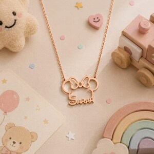 Op de afbeelding: Ros&eacute;gouden ketting met een Minnie Mouse-ontwerp en de naam "SARAH". De ketting wordt getoond met een pluche ster, een lachend hart, een houten trein en een regenboog.