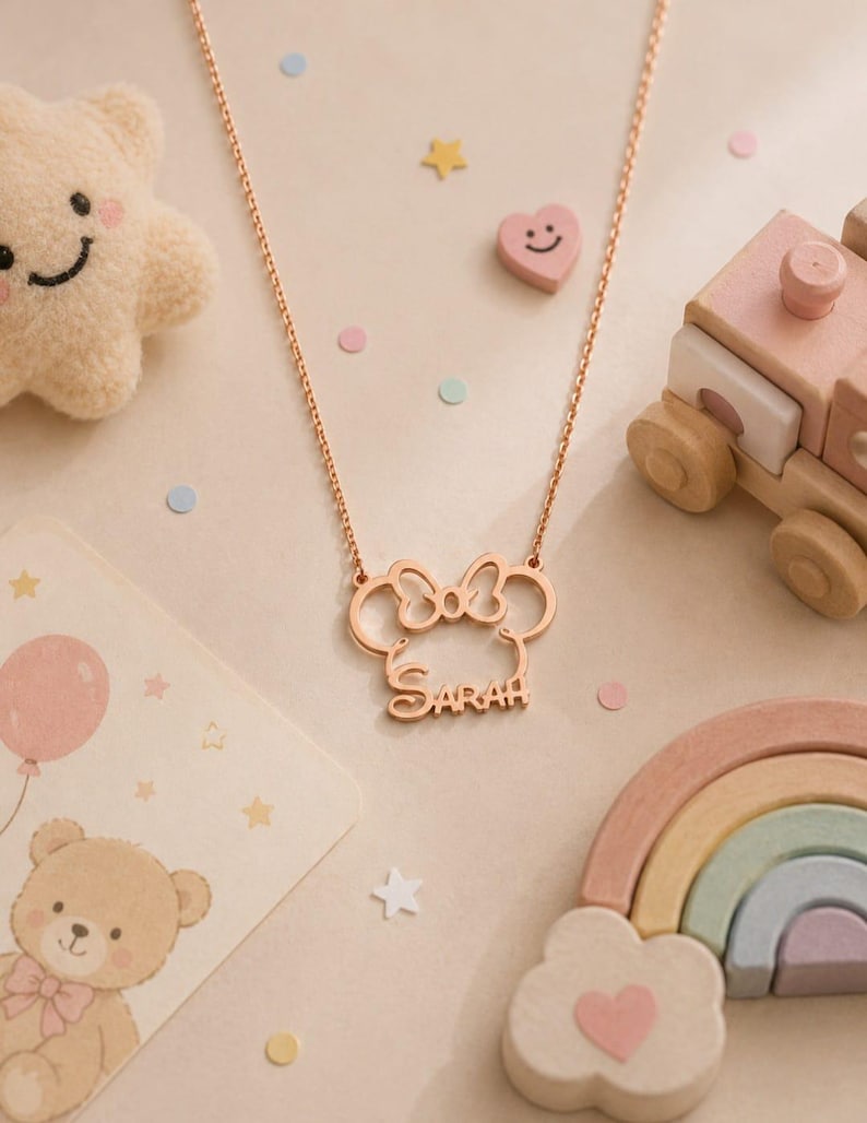 Op de afbeelding: Ros&eacute;gouden ketting met een Minnie Mouse-ontwerp en de naam "SARAH". De ketting wordt getoond met een pluche ster, een lachend hart, een houten trein en een regenboog.