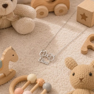 Op de afbeelding: Een zilveren ketting met de naam "Elena" in sierlijke letters, versierd met een strikvormig ontwerp. De ketting wordt getoond met kinderspeelgoed, waaronder een teddybeer, houten blokken en een speelgoedauto.