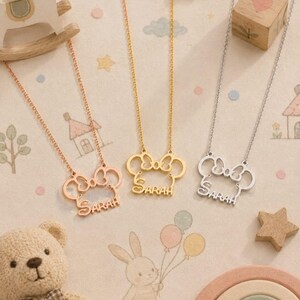 Pu&ograve; includere: Tre collane con ciondoli a forma di fiocco e il nome "SARAH" in corsivo. Le collane sono in oro rosa, oro e argento. Lo sfondo include un orsacchiotto, un cavallo a dondolo e un blocchetto di legno.