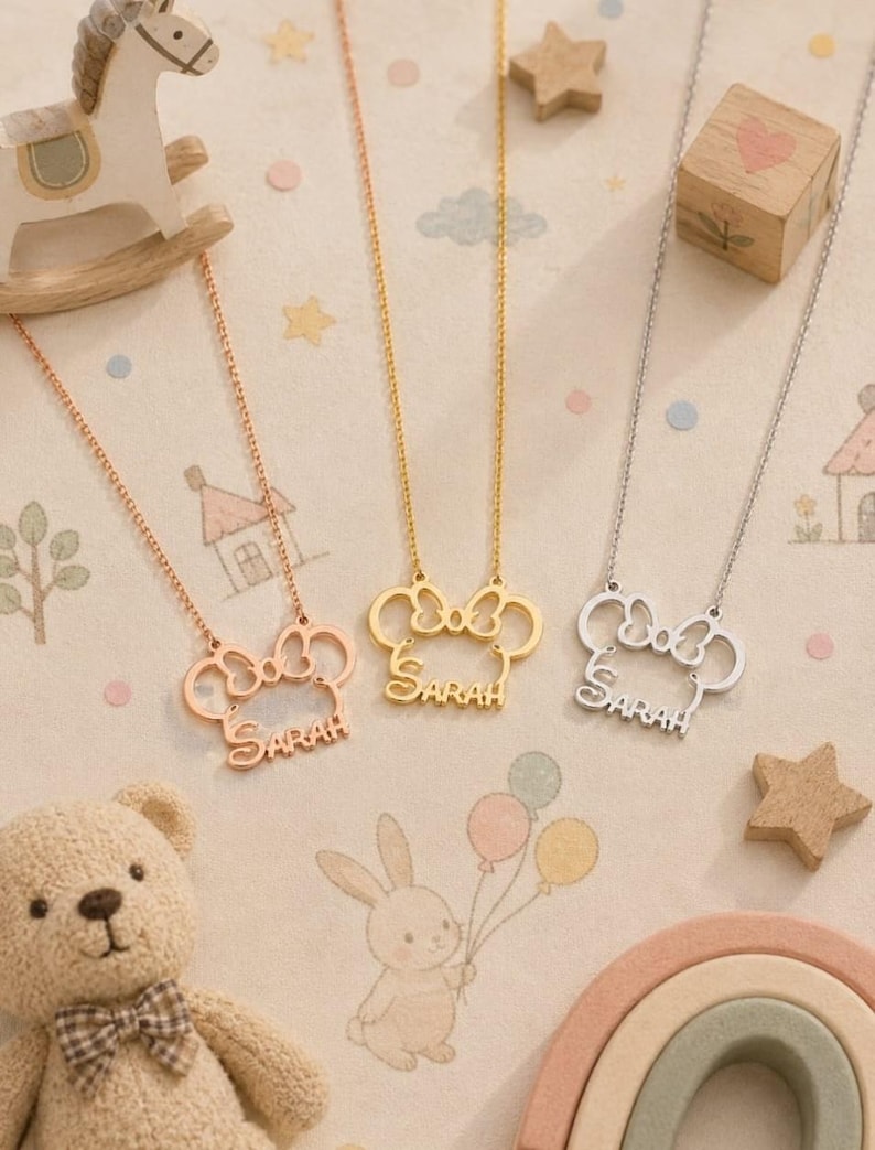 Pu&ograve; includere: Tre collane con ciondoli a forma di fiocco e il nome "SARAH" in corsivo. Le collane sono in oro rosa, oro e argento. Lo sfondo include un orsacchiotto, un cavallo a dondolo e un blocchetto di legno.