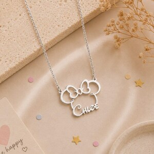 Op de afbeelding: Een zilverkleurige ketting met een Minnie Mouse-ge&iuml;nspireerd ontwerp. De hanger heeft een strik en de naam "Chloe". De ketting ligt op een beige oppervlak met decoratieve elementen.