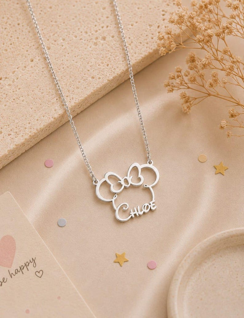 Op de afbeelding: Een zilverkleurige ketting met een Minnie Mouse-ge&iuml;nspireerd ontwerp. De hanger heeft een strik en de naam "Chloe". De ketting ligt op een beige oppervlak met decoratieve elementen.