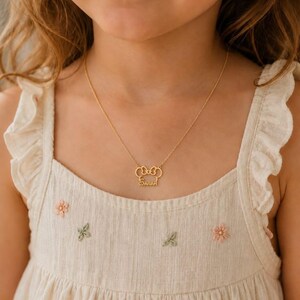 Pu&ograve; includere: Una collana color oro con un ciondolo a forma di fiocco con il nome "Sarah" inciso sotto. La collana &egrave; indossata da una persona che indossa un vestito color crema con ricami floreali.