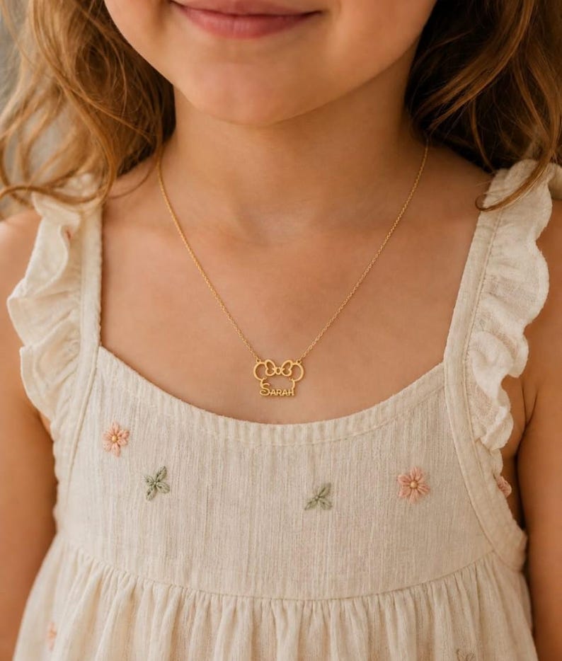 Pu&ograve; includere: Una collana color oro con un ciondolo a forma di fiocco con il nome "Sarah" inciso sotto. La collana &egrave; indossata da una persona che indossa un vestito color crema con ricami floreali.