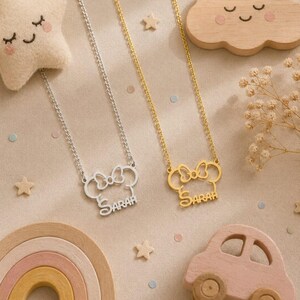 Pu&ograve; includere: Due collane con design di Minnie Mouse, una argentata e una dorata, ciascuna con il nome "SARAH" inciso. Le collane sono esposte con giocattoli di legno, tra cui una stella, una nuvola, un arcobaleno e un'auto, su una superficie beige.