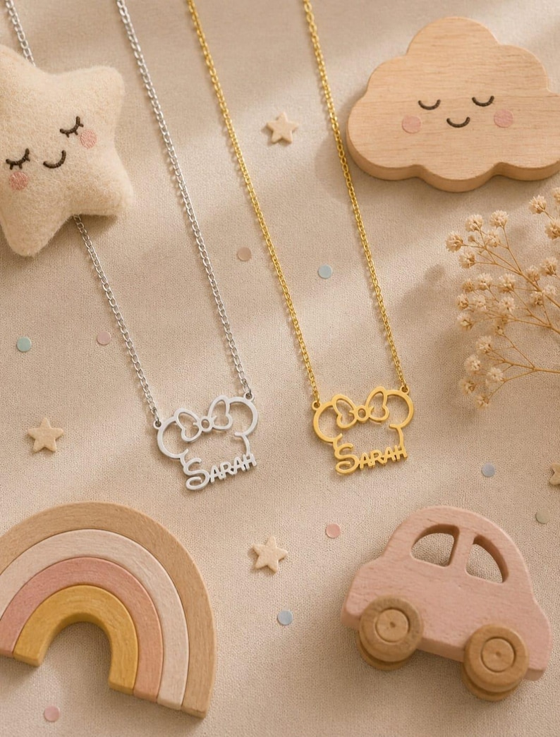 Pu&ograve; includere: Due collane con design di Minnie Mouse, una argentata e una dorata, ciascuna con il nome "SARAH" inciso. Le collane sono esposte con giocattoli di legno, tra cui una stella, una nuvola, un arcobaleno e un'auto, su una superficie beige.