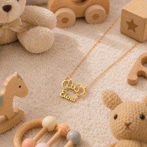 Op de afbeelding: Een goudkleurige ketting met de naam "Elena" in een sierlijk lettertype, omlijst door een strikvorm. De ketting wordt getoond met kinderspeelgoed, waaronder een teddybeer, houten blokken en een schommelpaard.