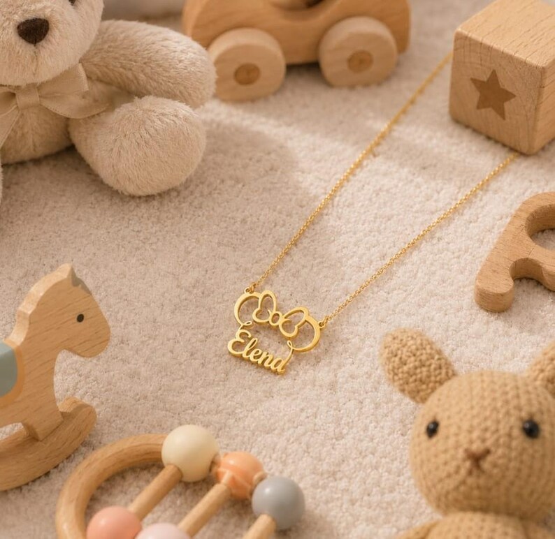 Op de afbeelding: Een goudkleurige ketting met de naam "Elena" in een sierlijk lettertype, omlijst door een strikvorm. De ketting wordt getoond met kinderspeelgoed, waaronder een teddybeer, houten blokken en een schommelpaard.