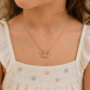 Op de afbeelding: Een goudkleurige ketting met een strikvormige hanger en de naam "Sarah" gegraveerd. De ketting wordt gedragen over een witte jurk met bloemenborduurwerk.
