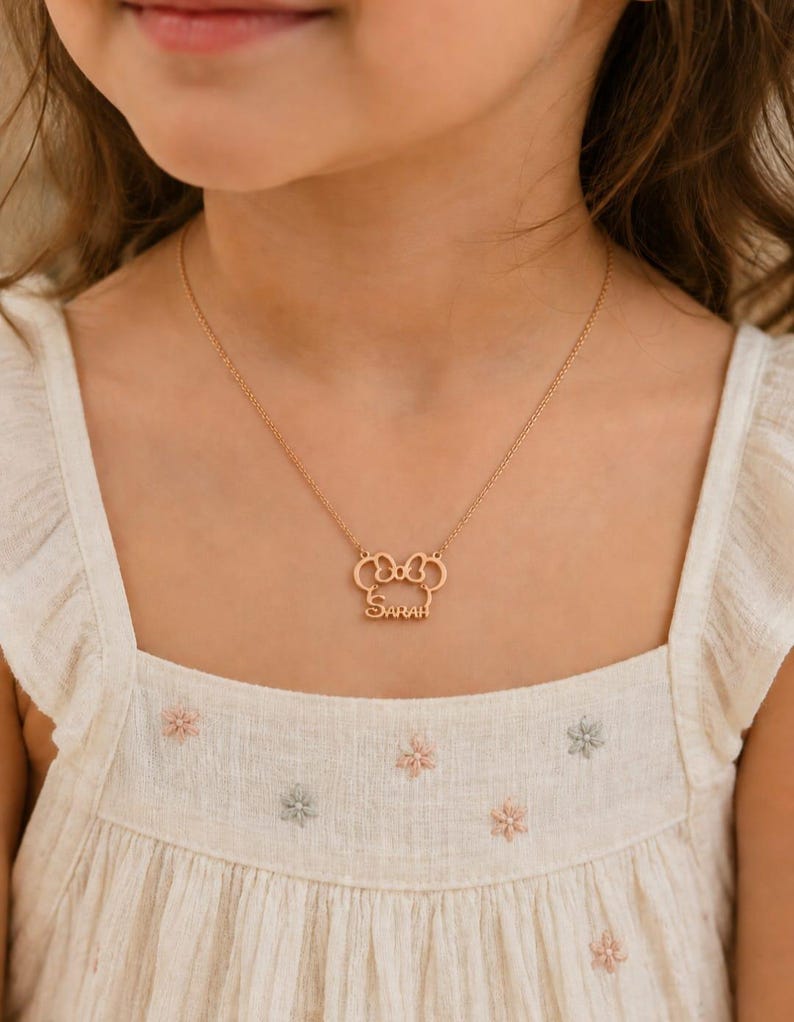 Op de afbeelding: Een goudkleurige ketting met een strikvormige hanger en de naam "Sarah" gegraveerd. De ketting wordt gedragen over een witte jurk met bloemenborduurwerk.