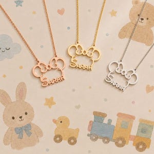 Op de afbeelding: Vier kettingen met de naam "SARAH" in een scriptlettertype. De hangers hebben de vorm van een strik met een muisoren-ontwerp. De kettingen zijn in ros&eacute;goud, goud en zilver. De achtergrond heeft een pastel babymotief met cartoon dieren.
